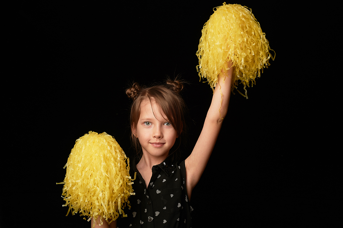 A girl of 6 years, on a black background. A cheerleader with yellow pumpons.Sport and energy.